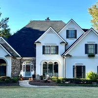 Large white two-story house with dark shutters, stone accents, and a manicured front lawn on a sunny day