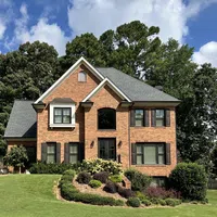 Two-story brick house with black shutters and landscaped front yard under blue sky with clouds.