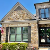 Stone and brick house exterior with black shutters, large windows, double front doors with green wreaths, and landscaping.