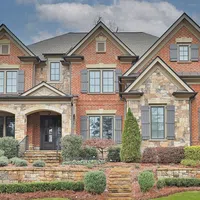 Large two-story brick and stone house with multiple windows, shutters, and landscaped front yard under blue sky.