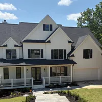 Two-story white house with black shutters, front porch, stone walkway, and surrounding greenery under blue sky.