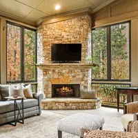 Cozy sunroom with stone fireplace, gray sofa, wicker chairs, large windows showing autumn foliage outdoors.