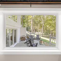 View through a large window of a wooden deck with Adirondack chairs and forest in the background.