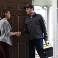 A service technician greets a homeowner at the front door, offering assistance and smiling.