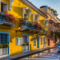 Colorful yellow buildings with flower-filled balconies lining a sunny street with a blue tuk-tuk parked nearby.