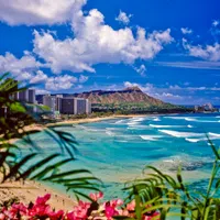 Waikiki Beach with turquoise water, surfers, city skyline, Diamond Head crater, and tropical foliage under a blue sky.