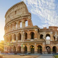 Sunrise over the ancient Colosseum in Rome with clear blue sky and surrounding greenery.