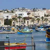 Colorful fishing boats docked in a Mediterranean harbor with stone buildings and palm trees under clear blue sky.