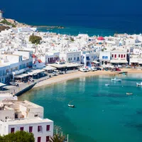 Aerial view of Mykonos town with white buildings, blue sea, boats, and windmills under a clear sky.