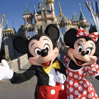Mickey and Minnie Mouse in front of Sleeping Beauty Castle at Disneyland, smiling and waving.