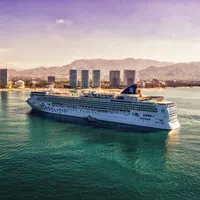 Cruise ship sailing near coastal city with high-rise buildings and mountains under a purple sky.