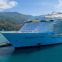 Large Symphony of the Seas cruise ship docked near lush green mountains under partly cloudy sky.