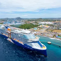 Large Celebrity Beyond cruise ship docked near tropical coastline with clear blue waters and city landscape in background