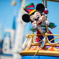 Mickey Mouse in colorful bandleader costume waving from a decorated float at a Disney castle parade under blue sky