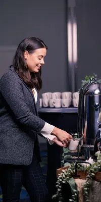 Woman in gray blazer pouring coffee from a large shiny urn at a coffee station with cups in background.