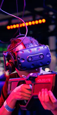 Person wearing a VR headset and holding a futuristic gaming gun in a neon-lit virtual reality arcade.