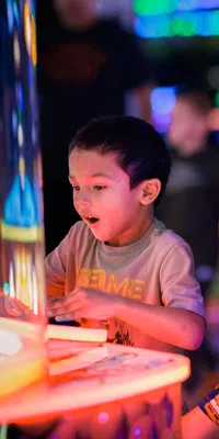 Young boy excitedly playing arcade game while woman watches and smiles in colorful gaming arcade environment