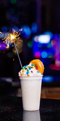 Milkshake with whipped cream, colorful candy, a donut, and a lit sparkler against a blurred night background.