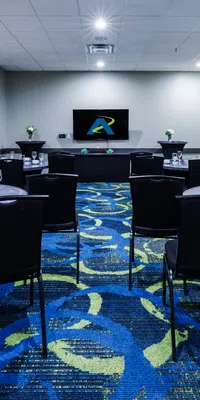 Conference room with round tables, black chairs, patterned blue carpet, and a screen at the front with logo.