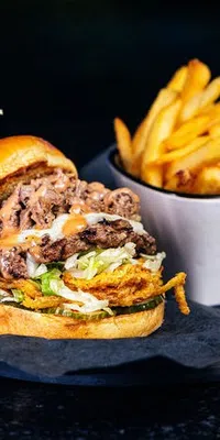 Juicy burger with beef, lettuce, sauce, and fries in a cup on a dark plate with a drink in the background