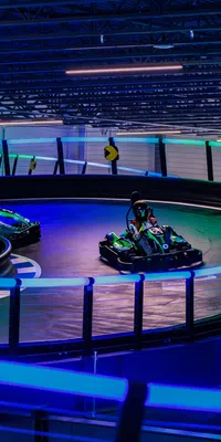 Indoor go-kart racing track with neon lights and racers navigating a sharp curve in dim lighting.