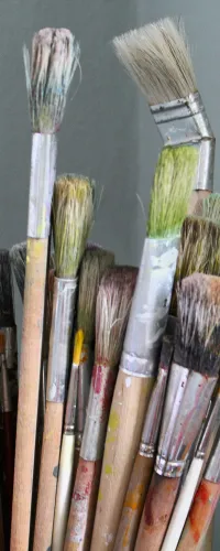 Assorted used paintbrushes with various bristle shapes and colors in a container against a gray wall background