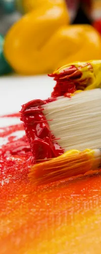 Close-up of paintbrushes with red and orange paint blending on a canvas, surrounded by colorful paint blobs.