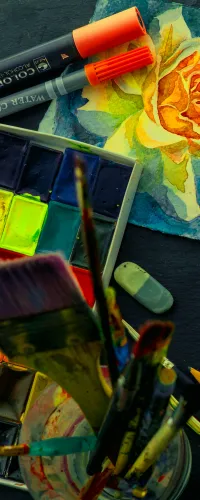 Watercolor painting supplies including brushes, pencils, watercolor palette, and a painted yellow rose on paper.