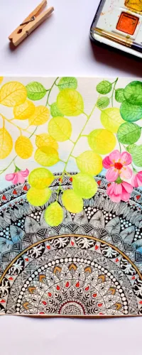 Watercolor painting of mandala and colorful flowers surrounded by paint set, cup of tea, and wooden clips on white surface