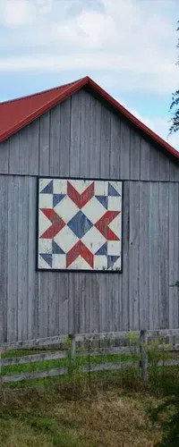 Gray wooden barn with a red roof featuring a red, white, and blue geometric quilt pattern on its side, surrounded by greenery.
