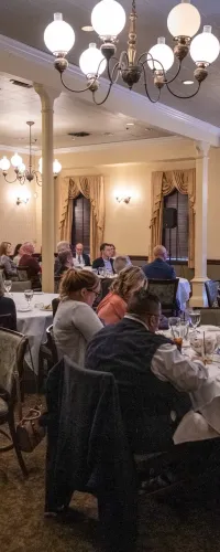 People attending a banquet-style meeting in an elegant room with chandeliers and a presenter at a screen.