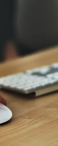 Hand holding a pen and using a white computer mouse next to a keyboard and monitor on a wooden desk