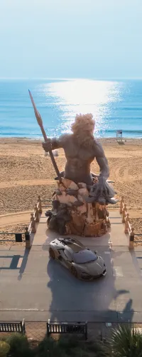 Statue of Poseidon with trident on beach promenade beside luxury sports car under clear blue sky.