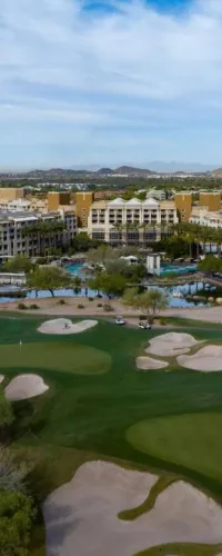 Aerial view of a resort with a large hotel, swimming pools, and a golf course surrounded by desert landscape.