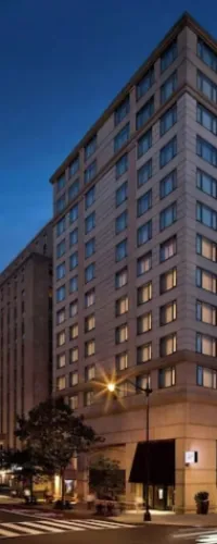 Modern multi-story hotel building illuminated at dusk on a city street corner with trees and crosswalk.