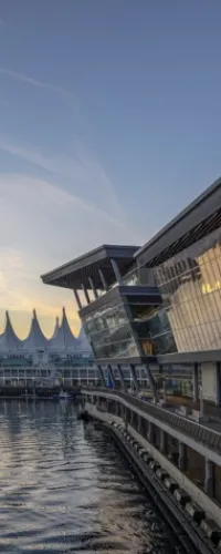 Modern waterfront building with glass facade reflecting sunset, adjacent to white pointed roof structures over water.