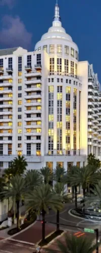 Illuminated Art Deco-style hotel building with palm trees and roundabout at dusk under a clear sky