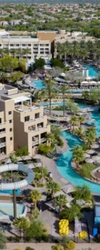 Aerial view of a resort with pools, water slides, palm trees, and surrounding buildings in a desert landscape.