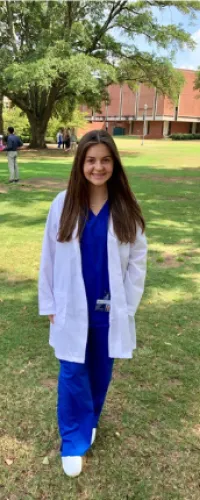 Smiling medical student in scrubs poses outdoors on campus during a sunny day.
