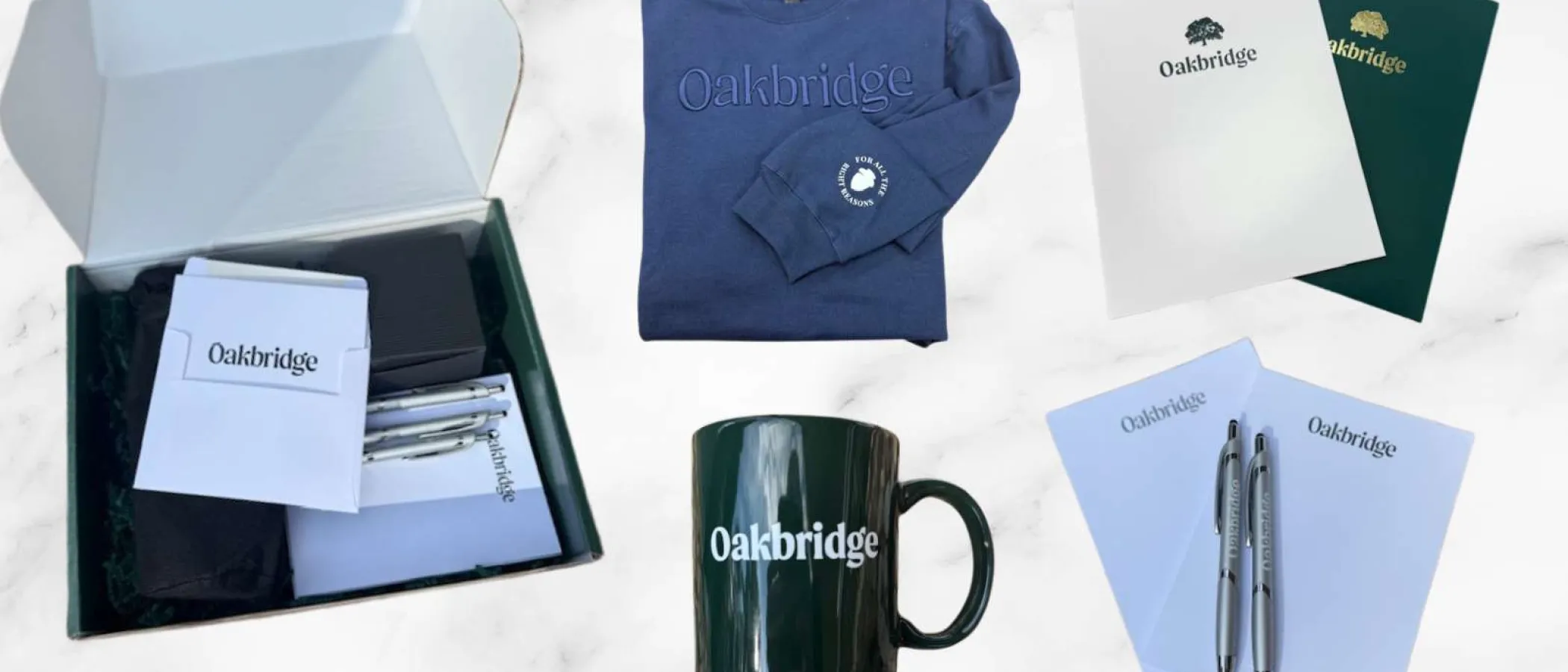 Oakbridge branded merchandise including a navy sweatshirt, black mug, pens, notebooks, and folders on a marble background.