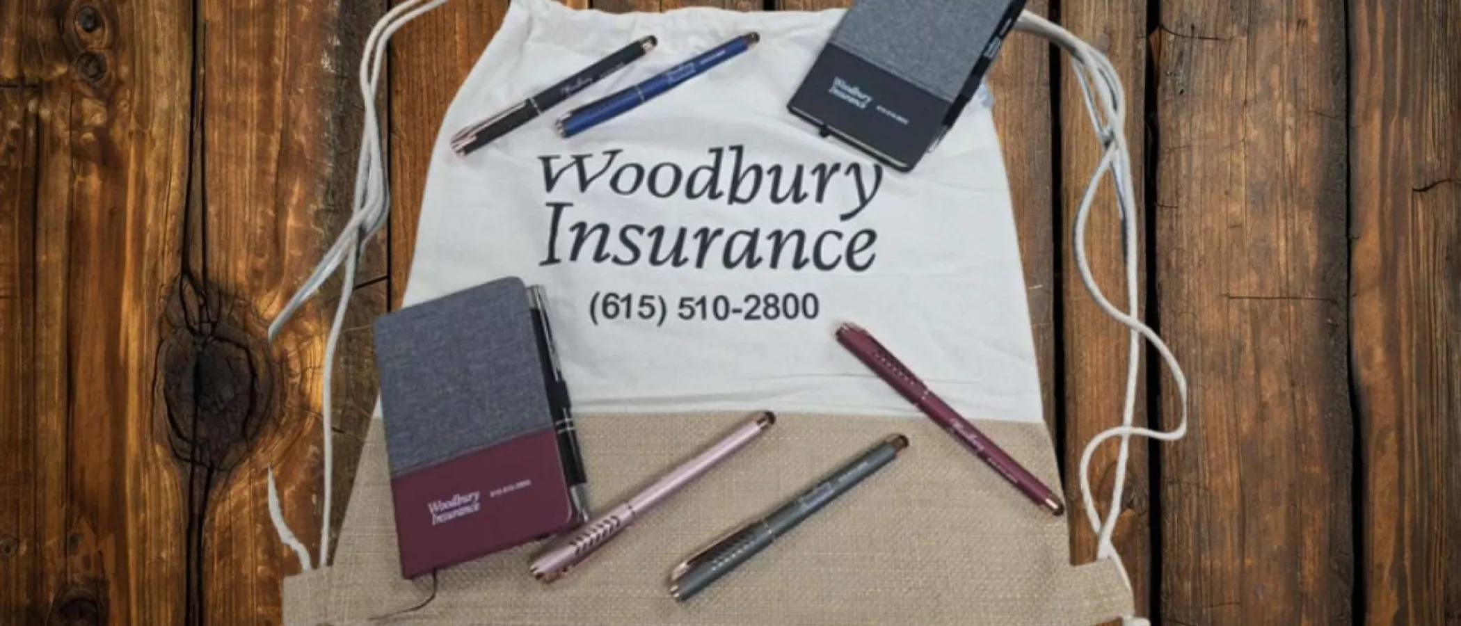 Woodbury Insurance drawstring bag with pens and notebooks on wooden surface showing phone number