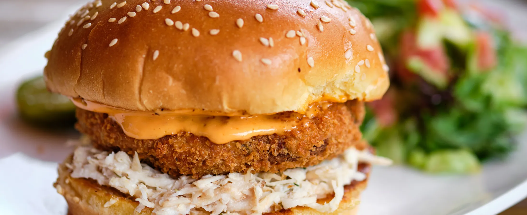 Close-up of a crispy fish sandwich with shredded fish, spicy sauce, and a sesame seed bun on a plate with salad.
