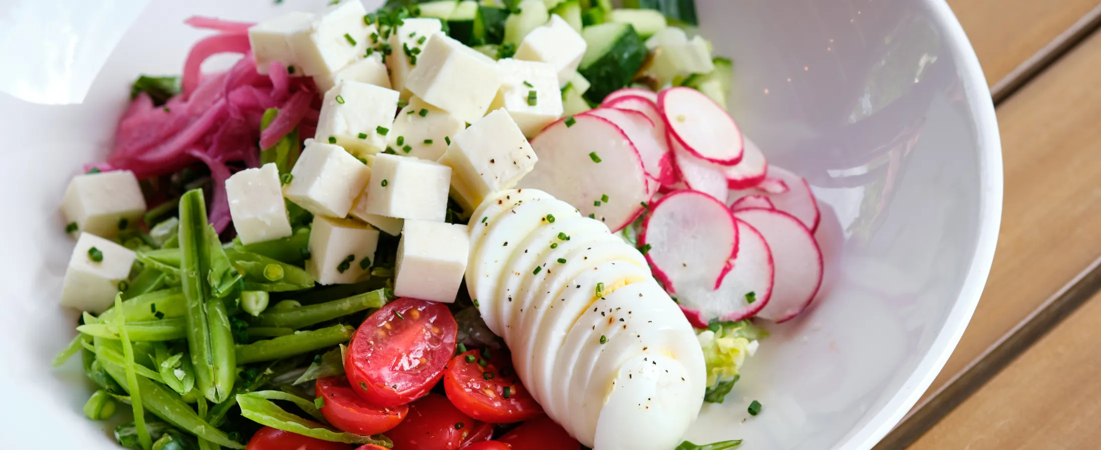 Fresh vegetable salad with cherry tomatoes, radishes, cucumber, tofu, and sliced boiled egg in white bowl.