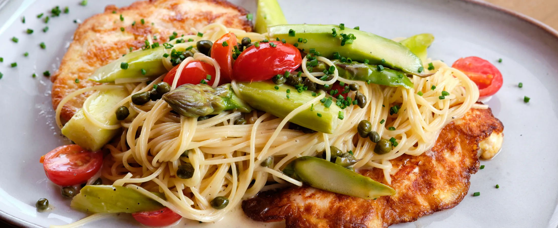 Grilled chicken breast served with angel hair pasta, asparagus, cherry tomatoes, and capers on a white plate.