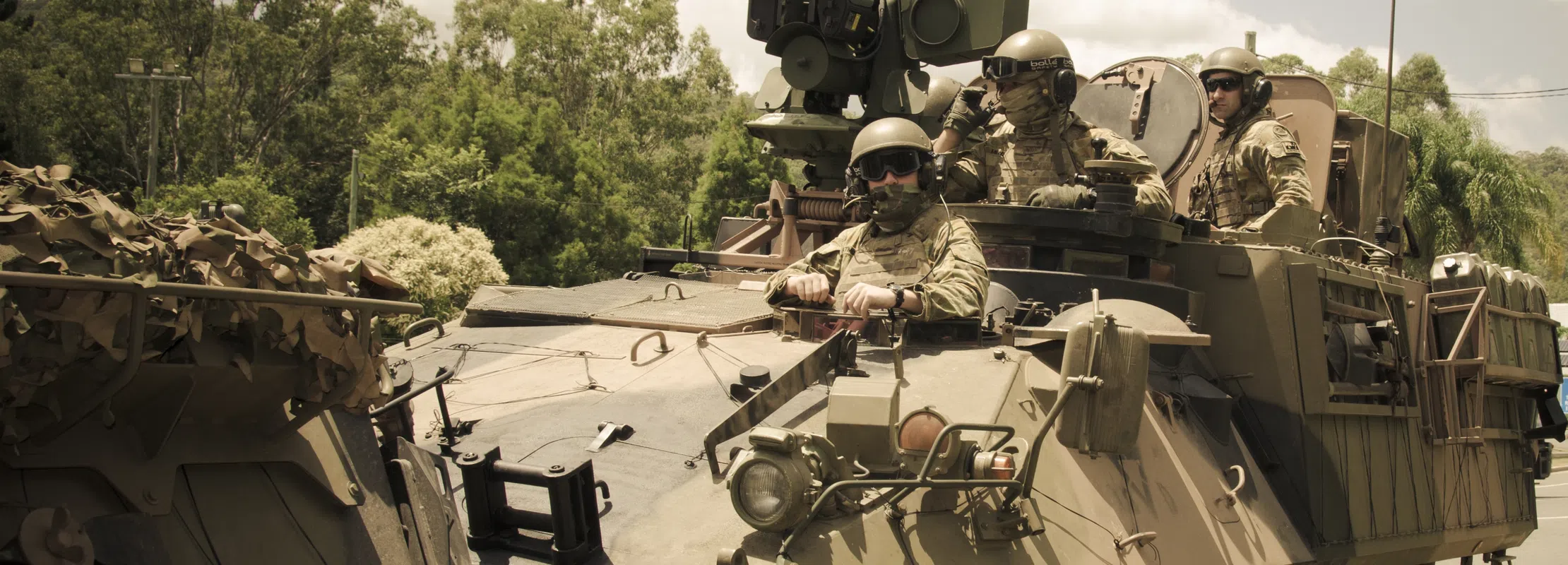 Military armored vehicle with soldiers wearing helmets and camouflage gear in a forested area under daylight.