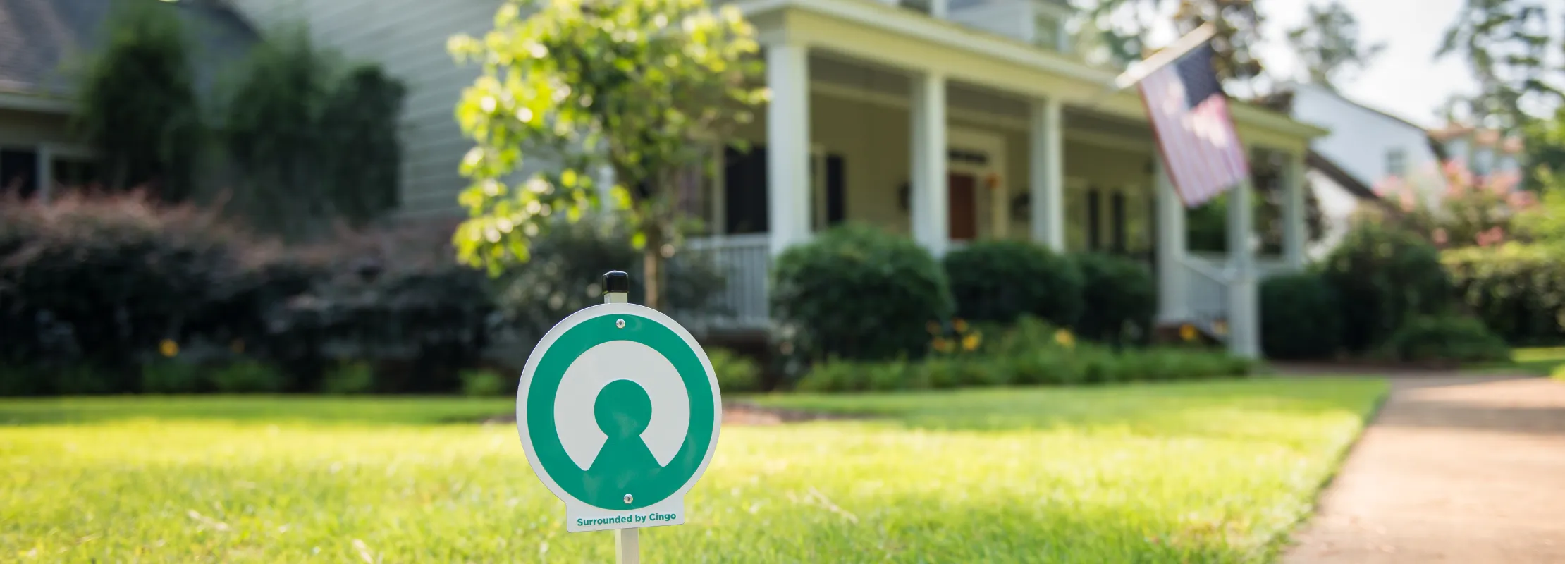 Security sign with a green shield logo on a lawn in front of a suburban house with a porch and American flag.