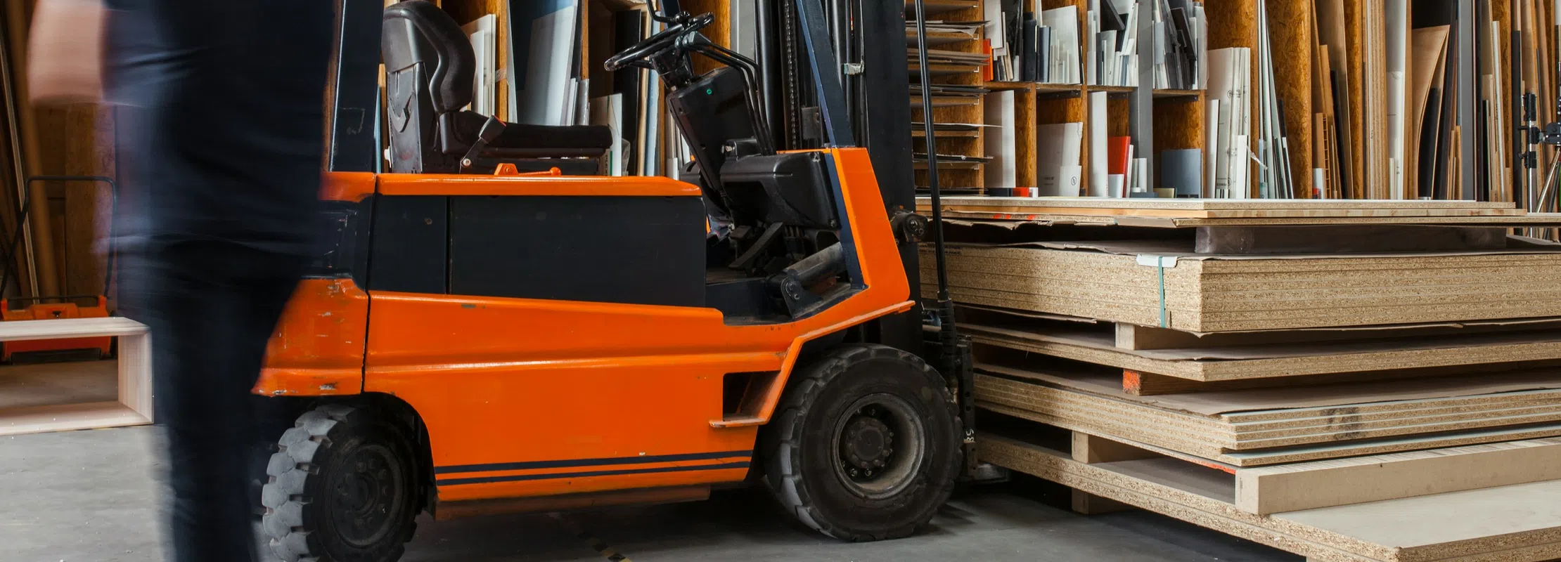 Orange forklift parked inside a warehouse with stacked wood panels and pallets in the background