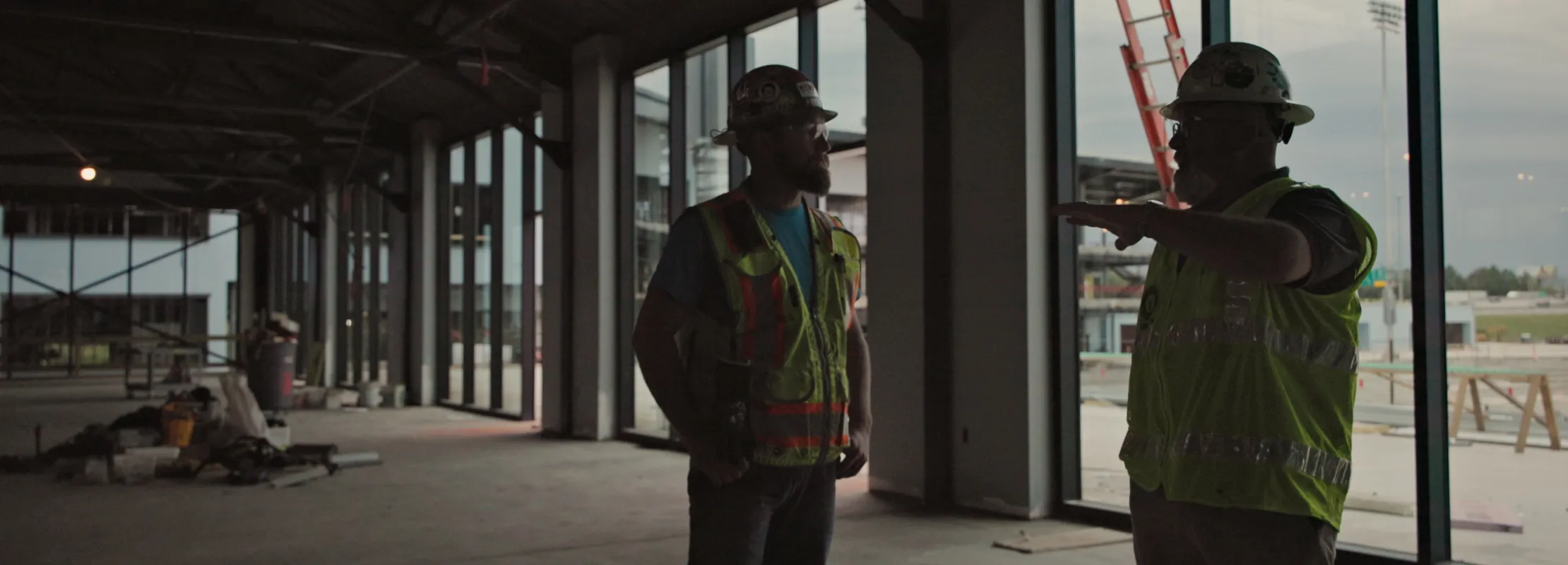 Two construction workers in safety gear discuss plans inside a large, unfinished building with large windows.