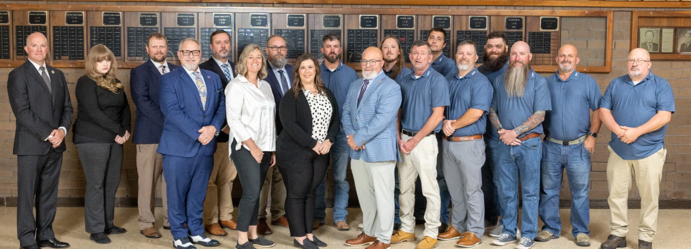 Group of 17 professionals posing indoors against a wall with portraits and plaques, dressed in business and casual attire.