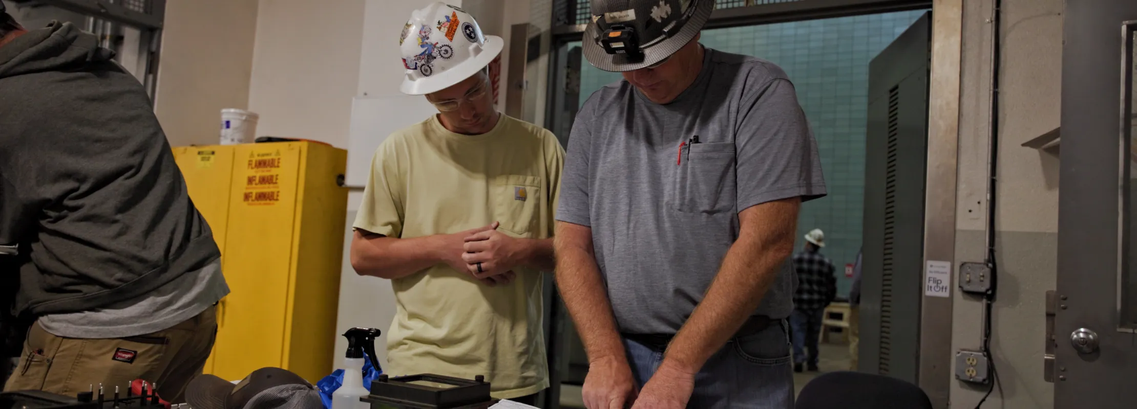 Two construction workers wearing helmets reviewing blueprints inside a worksite with equipment and tools.